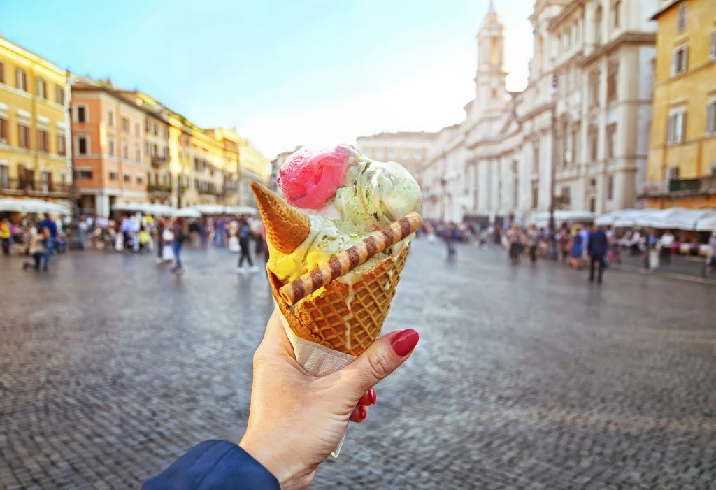A woman’s hand holding tri-colored ice cream cone with an Italian piazza in the background.