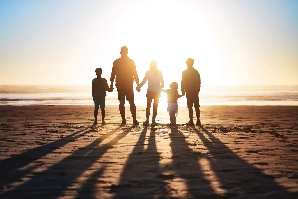 Silhouette of parents and 3 children holding hands on beach with sun casting shadows behind them.