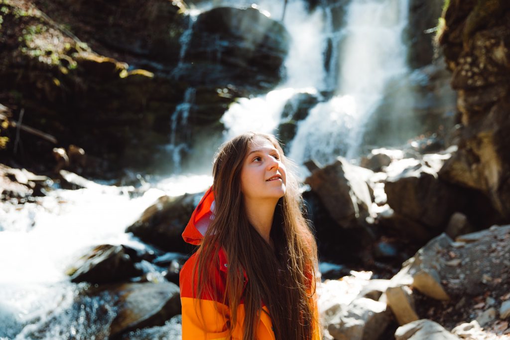Young woman wears red-orange rain coat that stands out against cool waterfall in background.