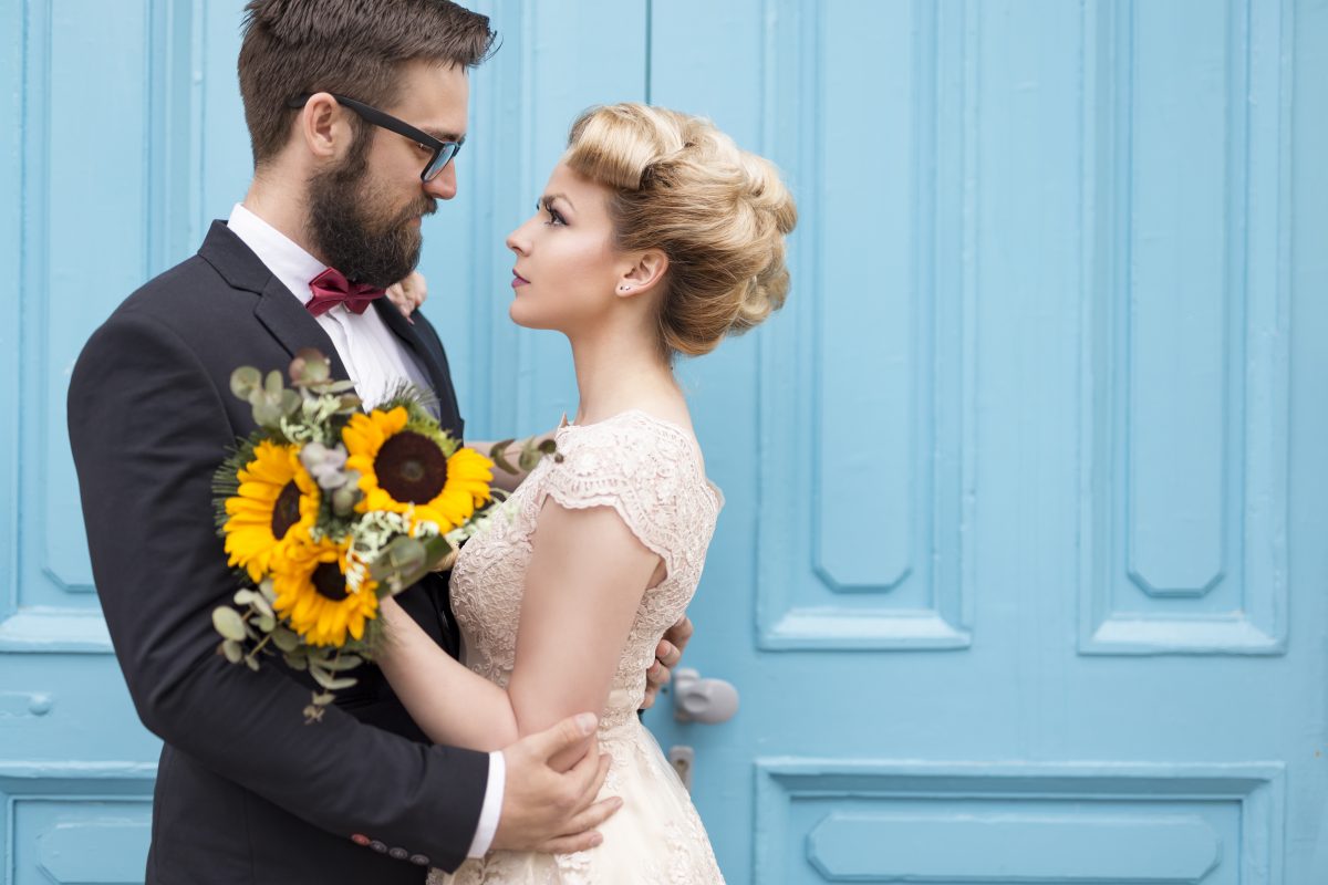 Newlyweds embrace in contrasting color photography, where sunflower bouquet contrasts blue doors.