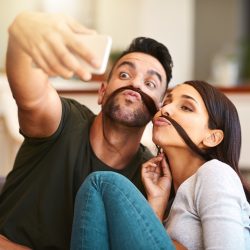 Young couple creates funny selfie ideas by wrapping woman’s hair around upper lip like a mustache.