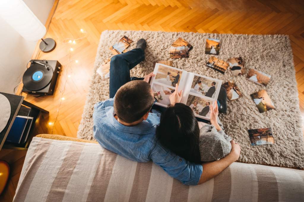 Couple on couch goes through wedding album photo book design with picture printouts on carpet.