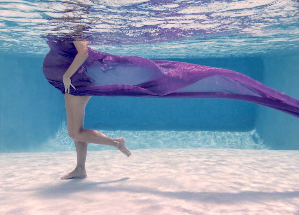 Pregnant woman in purple dress stands in a pool for an underwater photography shoot.