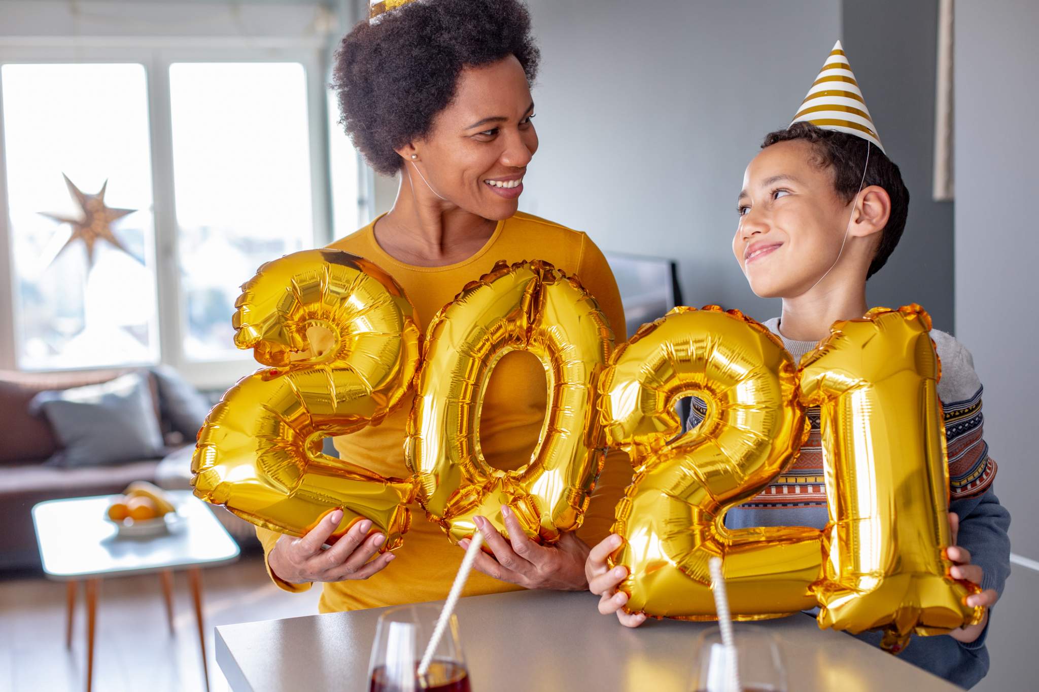Mom & young son wear festive hats and hold ‘2021’ balloons at home to take New Year’s Eve pictures.