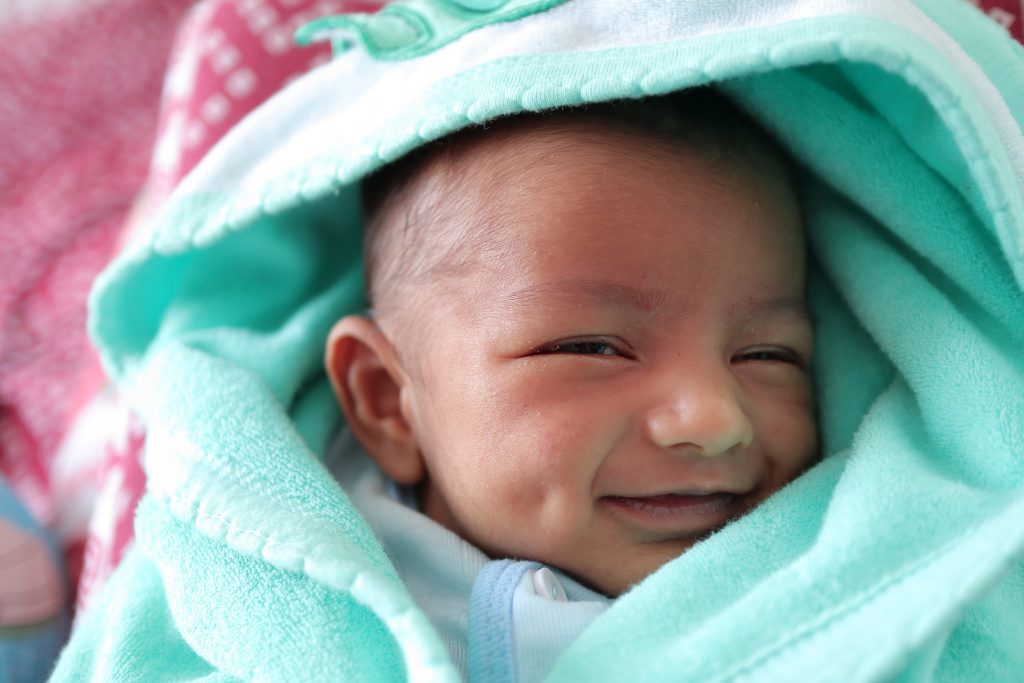 A smiling newborn baby with dimple in cheek wrapped in sea green colored towel with hood with eyes closed with selective focus on front eye