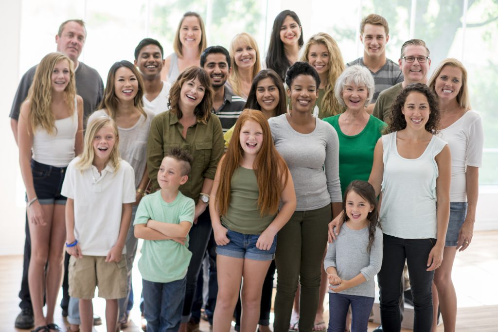 A multi-ethnic group of multiple generations are standing together for a family reunion. They are happily laughing and smiling while looking at the camera.