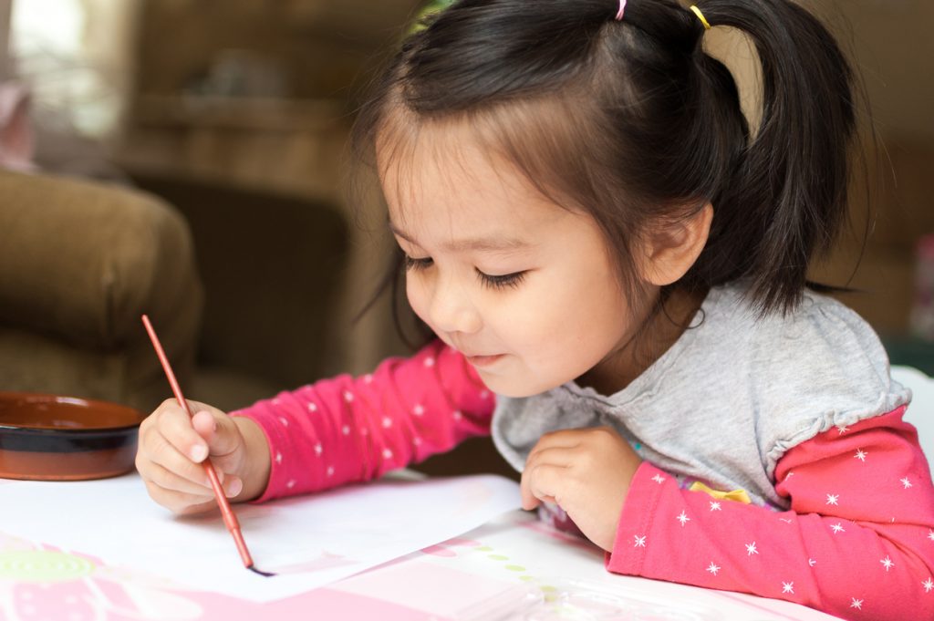A girl holds a small paintbrush and is in the process of stroking the bristles over a piece of paper.