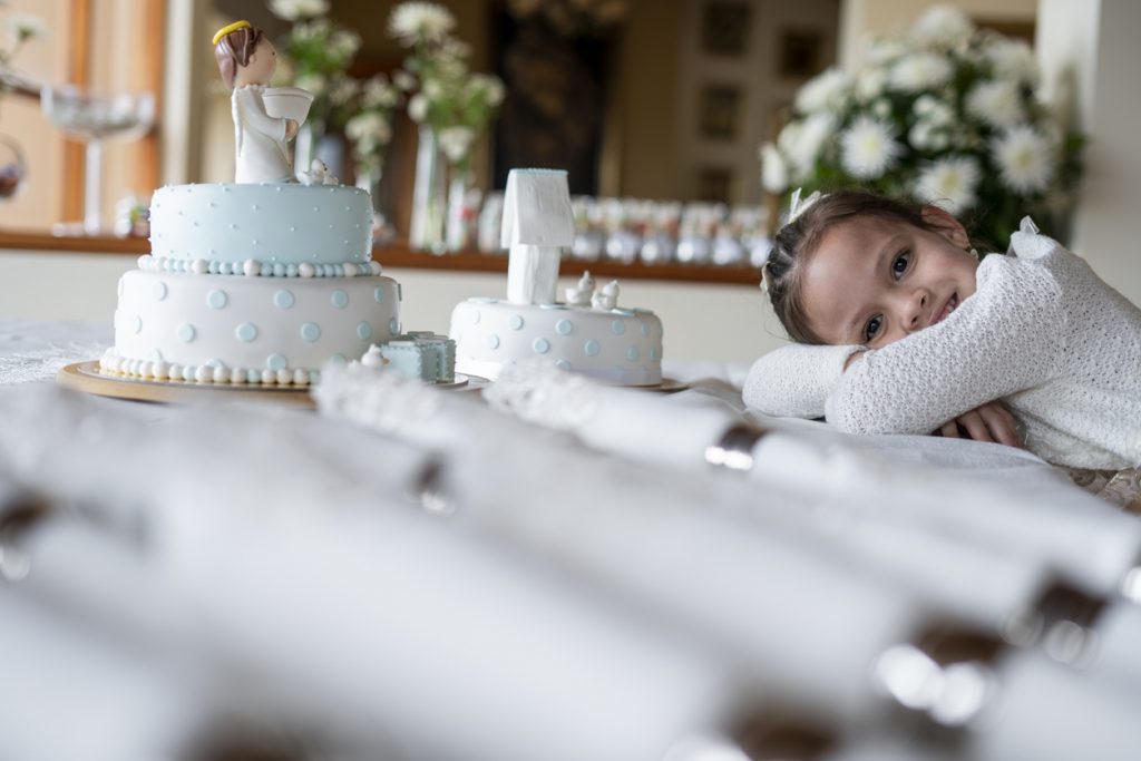 5-year-old Latin girl dressed in an elegant formal dress is inside the house where the celebration of the christening of a baby is held, she rests her head on the central table of the decoration where the cake and cutlery are located, she looks at the camera and smiles